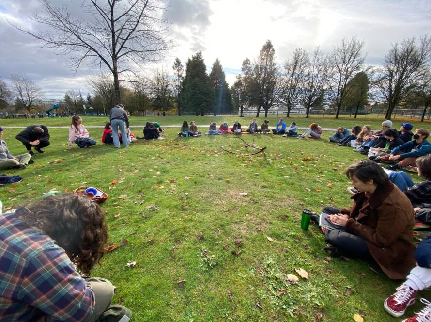 large circle of young people sitting in a park, green grass, overcast sky.