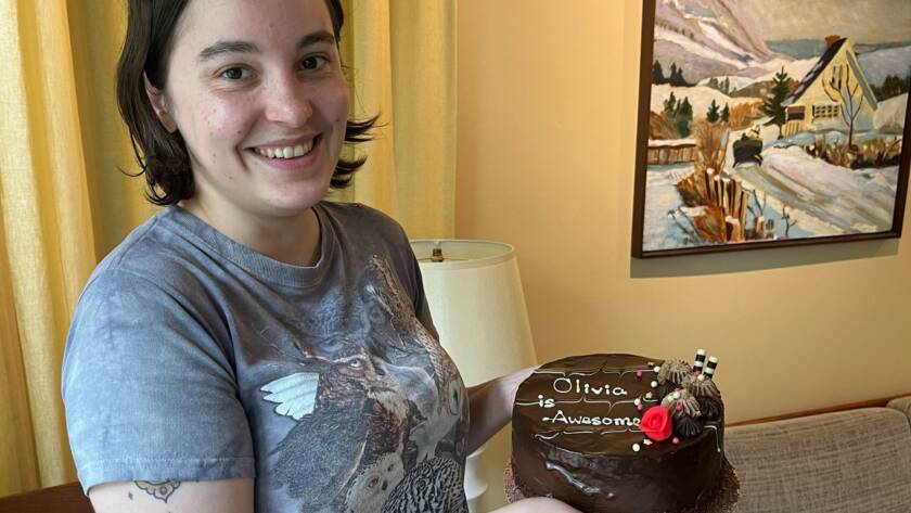 person with dark hair and grey t-shirt holding a chocolate cake that reads "Olivia is Awesome!"