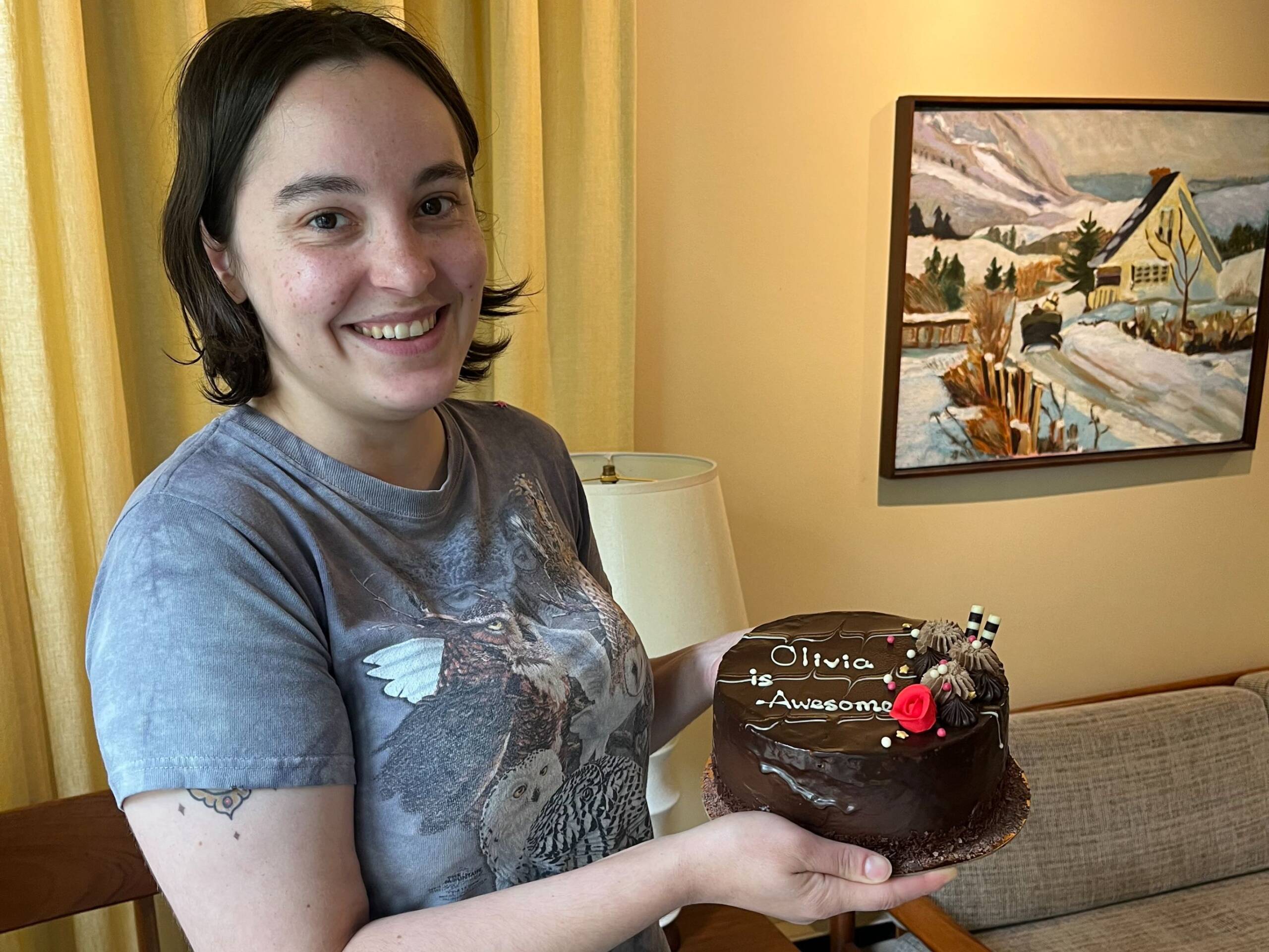 person with dark hair and grey t-shirt holding a chocolate cake that reads "Olivia is Awesome!"