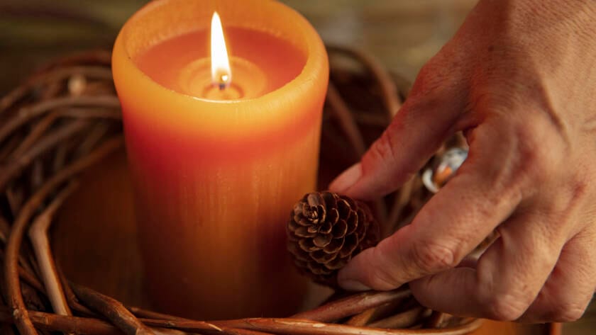 close-up on an orange candle surrounded by a vine wreath. A hand places a pine cone next to the candle.