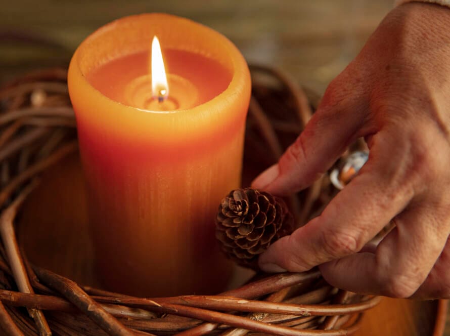 close-up on an orange candle surrounded by a vine wreath. A hand places a pine cone next to the candle.